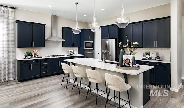 kitchen featuring a kitchen bar, a kitchen island with sink, a sink, appliances with stainless steel finishes, and wall chimney range hood