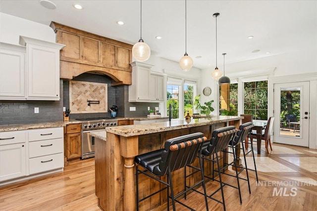 kitchen featuring high end range, hanging light fixtures, light stone counters, white cabinets, and a center island with sink