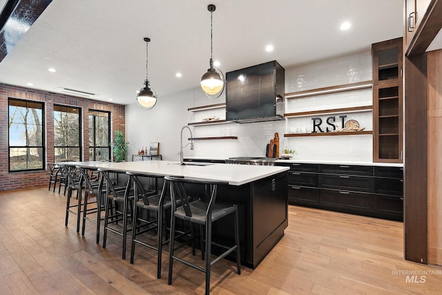 kitchen with wall chimney exhaust hood, sink, hanging light fixtures, an island with sink, and light hardwood / wood-style floors