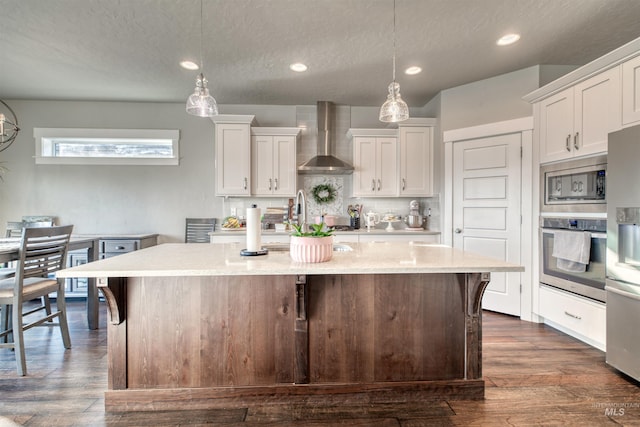 kitchen with hanging light fixtures, wall chimney exhaust hood, stainless steel appliances, white cabinets, and an island with sink