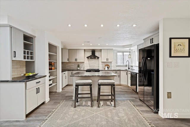 kitchen with wall chimney range hood, sink, black appliances, hardwood / wood-style flooring, and a center island