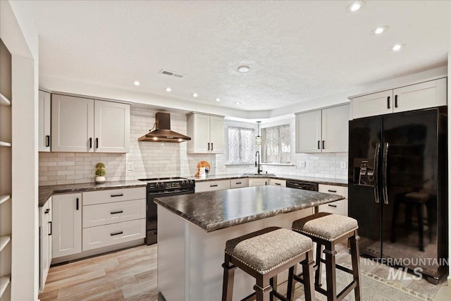 kitchen featuring sink, wall chimney range hood, a kitchen island, black appliances, and light wood-type flooring