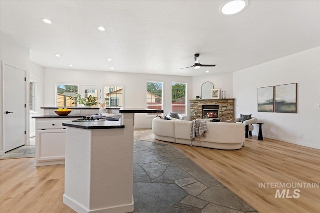 kitchen with dark countertops, a center island, a stone fireplace, light wood-style floors, and a ceiling fan