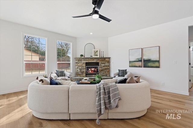 living room with a ceiling fan, a stone fireplace, wood finished floors, and baseboards