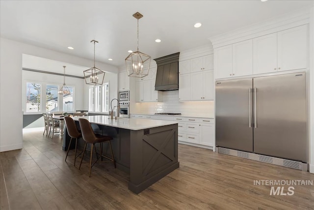 kitchen with decorative backsplash, an island with sink, dark wood-style floors, custom range hood, and built in appliances