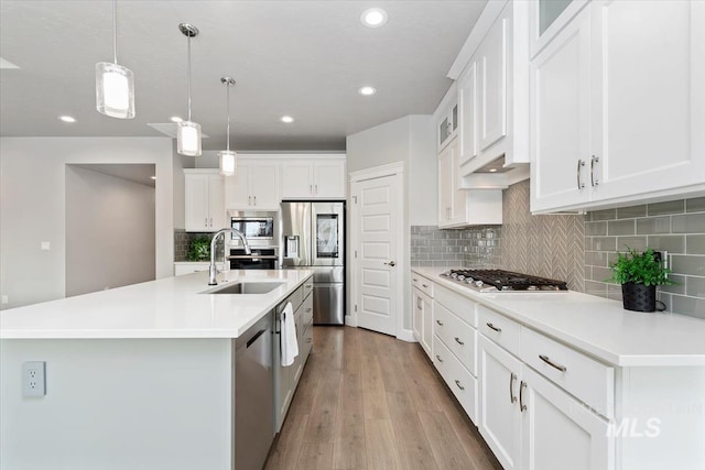 kitchen featuring a kitchen island with sink, wood finished floors, a sink, appliances with stainless steel finishes, and decorative light fixtures