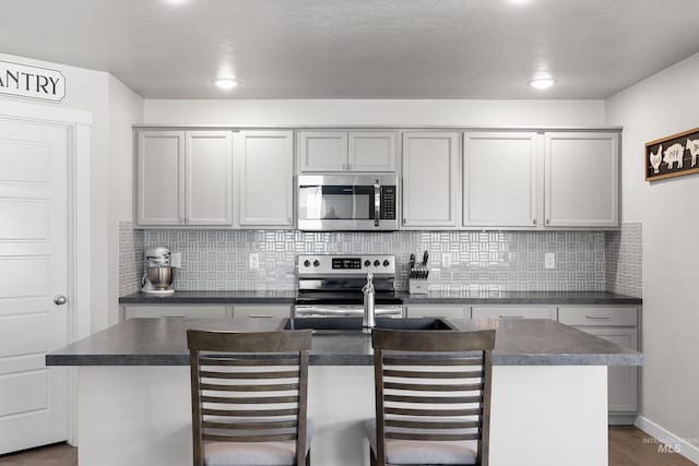 kitchen with dark wood finished floors, dark countertops, an island with sink, backsplash, and appliances with stainless steel finishes