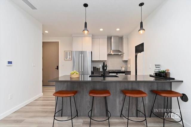 kitchen featuring freestanding refrigerator, dark countertops, a breakfast bar area, wall chimney exhaust hood, and a sink