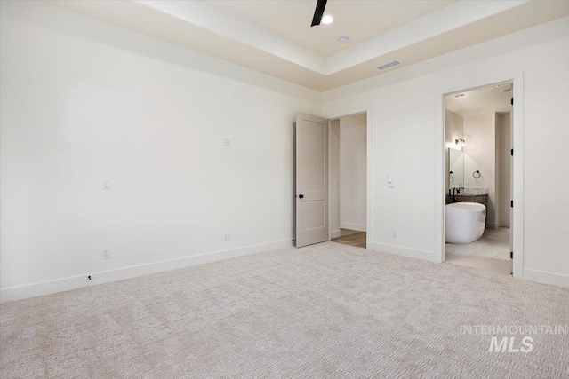 unfurnished bedroom featuring ceiling fan, ensuite bathroom, light colored carpet, and a tray ceiling