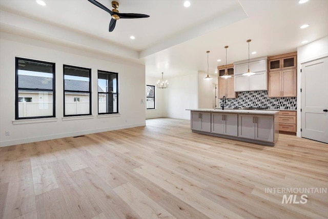 kitchen featuring decorative light fixtures, tasteful backsplash, a raised ceiling, a kitchen island with sink, and ceiling fan with notable chandelier