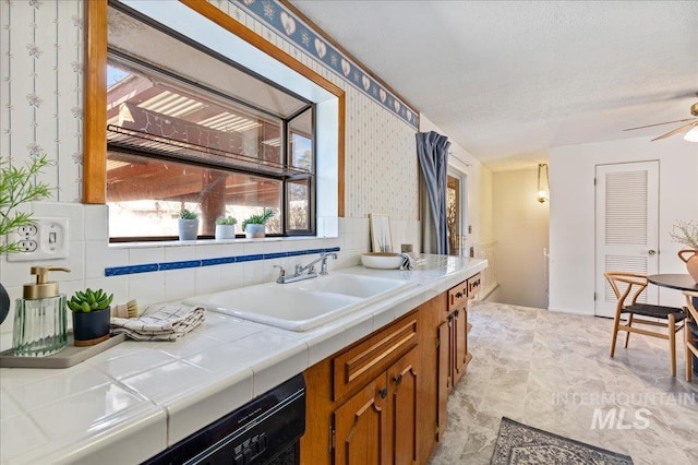 kitchen featuring brown cabinetry, wallpapered walls, ceiling fan, a sink, and tile counters