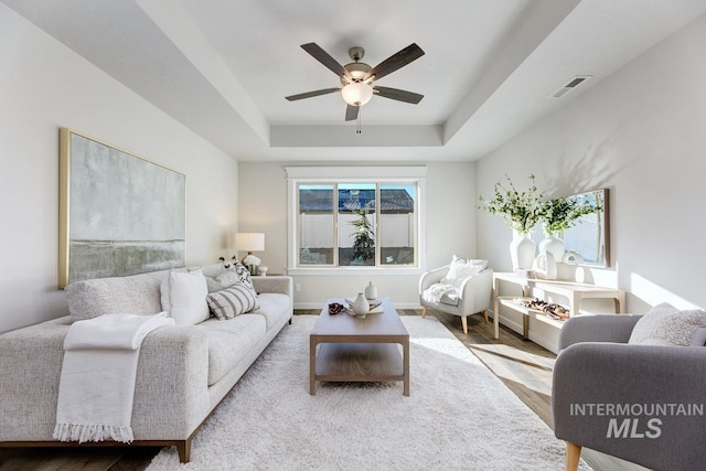 living area with a ceiling fan, visible vents, wood finished floors, and a raised ceiling