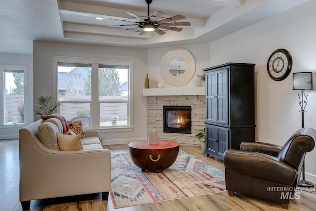 living room featuring a stone fireplace, wood finished floors, a ceiling fan, baseboards, and a raised ceiling