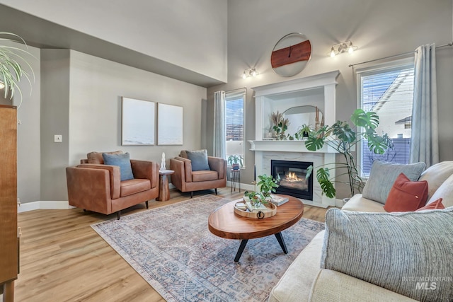 living area featuring light wood-style floors, a glass covered fireplace, a towering ceiling, and baseboards