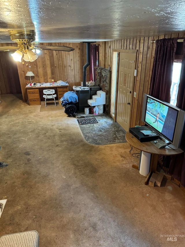 carpeted living room with a wood stove, ceiling fan, and wooden walls