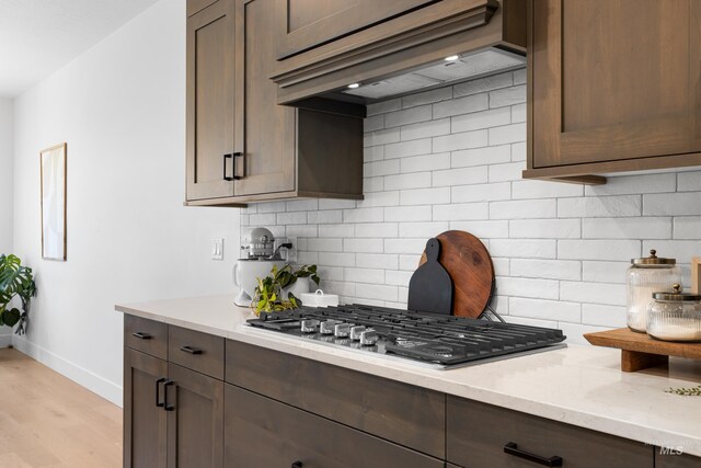 kitchen featuring stainless steel gas cooktop, custom exhaust hood, baseboards, decorative backsplash, and dark brown cabinets