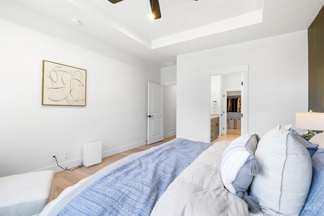 bedroom with a raised ceiling, light wood-style floors, ensuite bath, and a ceiling fan