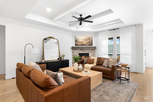living area with a ceiling fan, light wood-type flooring, a stone fireplace, a tray ceiling, and beamed ceiling