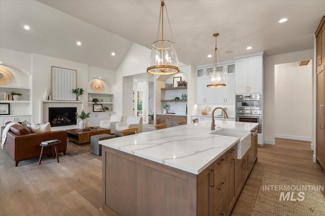 kitchen with light wood finished floors, a center island with sink, double oven, white cabinetry, and a sink