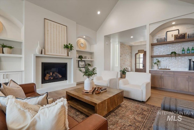 living room featuring built in shelves, wood finished floors, high vaulted ceiling, recessed lighting, and a glass covered fireplace