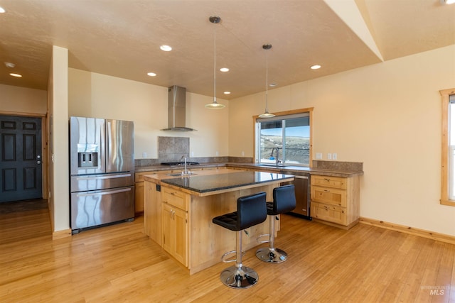 kitchen with light wood finished floors, stainless steel appliances, light brown cabinetry, wall chimney range hood, and an island with sink