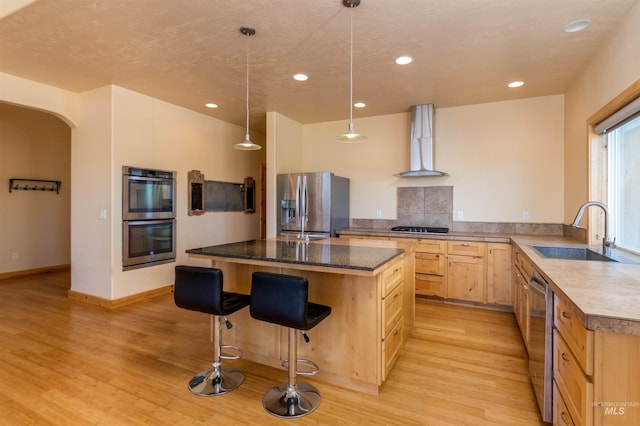 kitchen with arched walkways, stainless steel appliances, light brown cabinets, a sink, and wall chimney exhaust hood