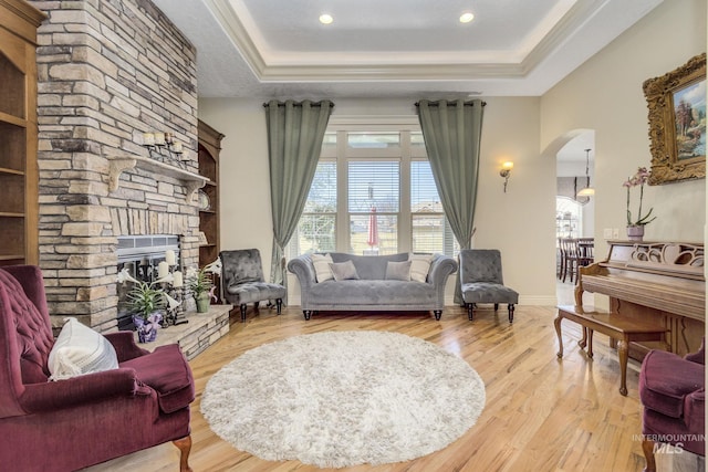 living room with arched walkways, a stone fireplace, a raised ceiling, and light wood-style flooring