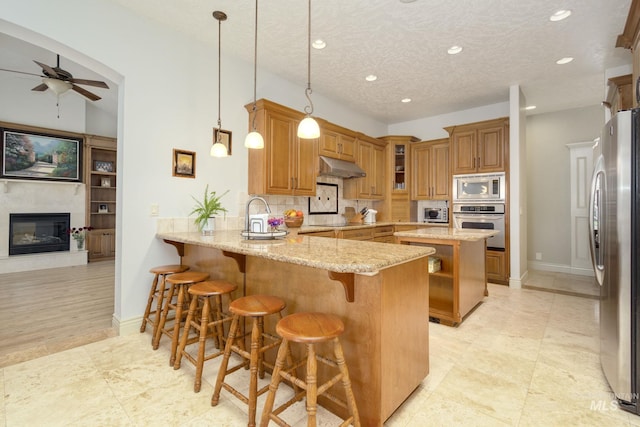 kitchen with light stone counters, under cabinet range hood, a peninsula, appliances with stainless steel finishes, and a glass covered fireplace