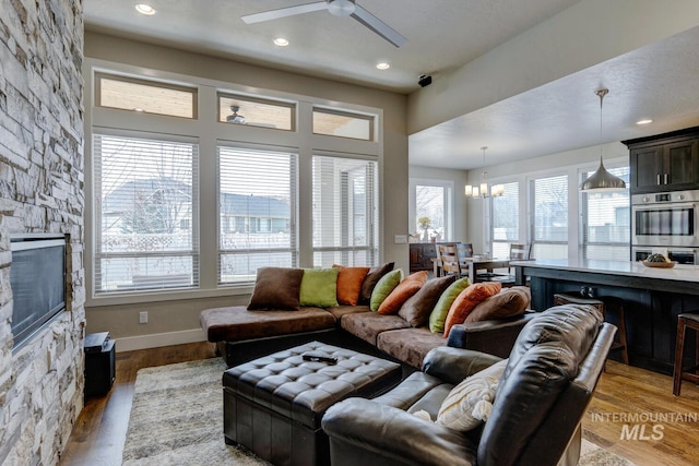 living room featuring a stone fireplace, recessed lighting, ceiling fan with notable chandelier, baseboards, and light wood-style floors