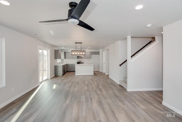 unfurnished living room featuring a sink, stairs, light wood finished floors, ceiling fan, and baseboards