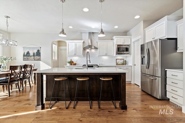 kitchen featuring wall chimney exhaust hood, white cabinetry, an island with sink, pendant lighting, and stainless steel appliances