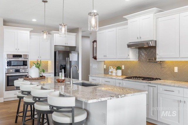 kitchen featuring stainless steel appliances, white cabinets, a kitchen island with sink, a sink, and under cabinet range hood