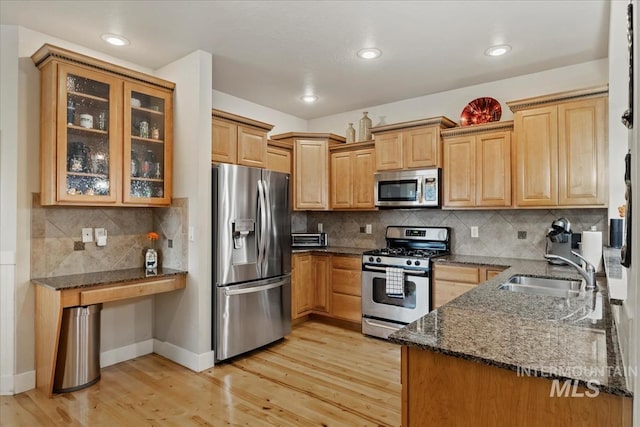 kitchen with light wood finished floors, glass insert cabinets, dark stone counters, appliances with stainless steel finishes, and a sink