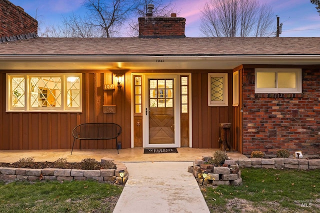 exterior entry at dusk featuring board and batten siding, a chimney, covered porch, and roof with shingles