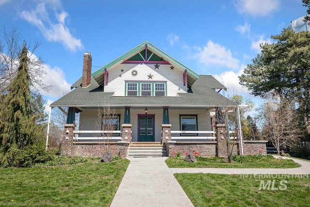 view of front facade featuring a shingled roof, a chimney, a front lawn, and covered porch