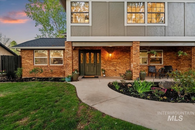 exterior entry at dusk featuring brick siding, fence, stucco siding, and a yard