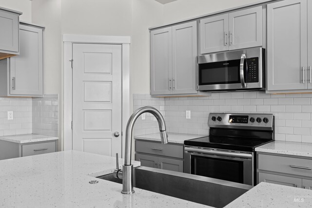 kitchen with appliances with stainless steel finishes, backsplash, light stone counters, gray cabinets, and a sink