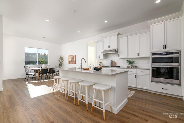 kitchen with hanging light fixtures, stainless steel appliances, white cabinets, and a center island with sink