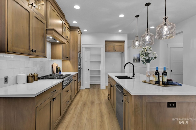 kitchen featuring a kitchen island with sink, light wood-style flooring, a sink, backsplash, and stainless steel appliances