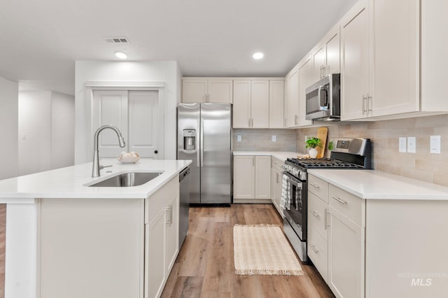 kitchen with sink, appliances with stainless steel finishes, a kitchen island with sink, white cabinets, and light wood-type flooring