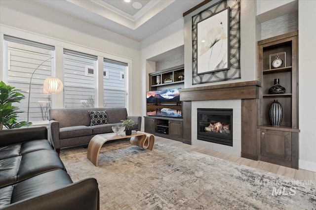 living room featuring a tile fireplace, a tray ceiling, crown molding, light wood-type flooring, and built in shelves