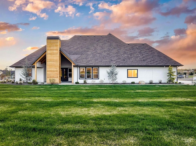 back of property featuring a chimney, stucco siding, a shingled roof, and a lawn