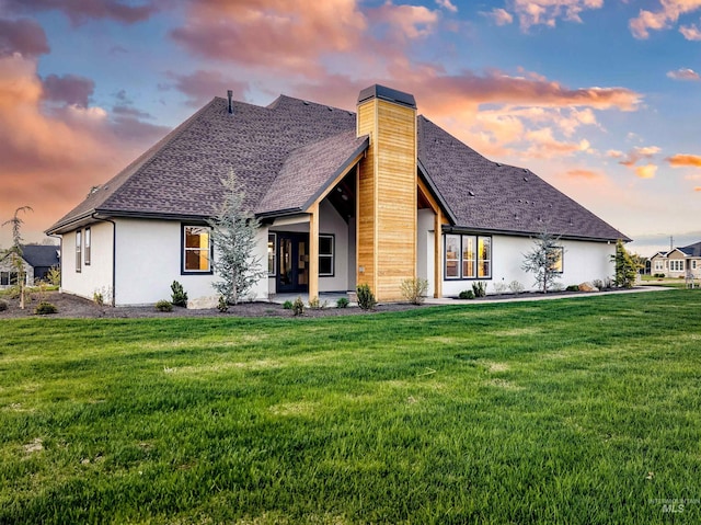 back of house with a lawn, a chimney, stucco siding, and roof with shingles