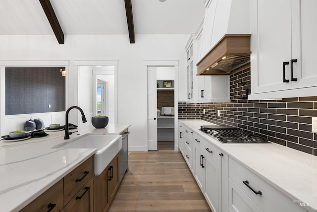 kitchen with a sink, beamed ceiling, light wood-style flooring, custom exhaust hood, and white cabinetry
