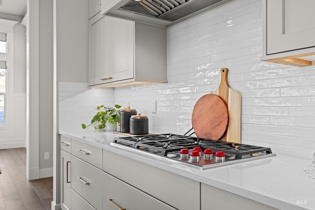 kitchen with custom exhaust hood, backsplash, stainless steel gas stovetop, light stone counters, and light hardwood / wood-style flooring