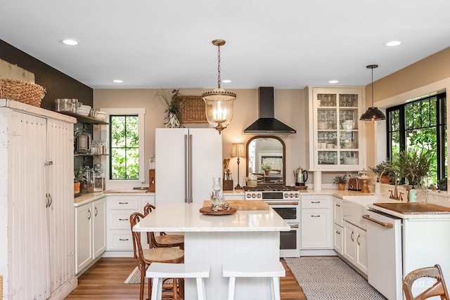 kitchen featuring pendant lighting, wall chimney range hood, white appliances, white cabinetry, and a kitchen island