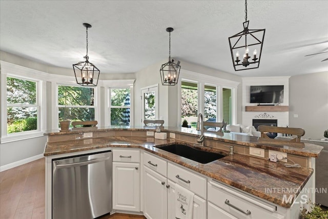 kitchen featuring a kitchen island with sink, sink, stainless steel dishwasher, a large fireplace, and white cabinetry