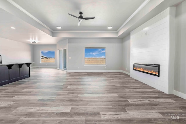 unfurnished living room featuring a raised ceiling, ceiling fan, light wood-type flooring, a fireplace, and crown molding