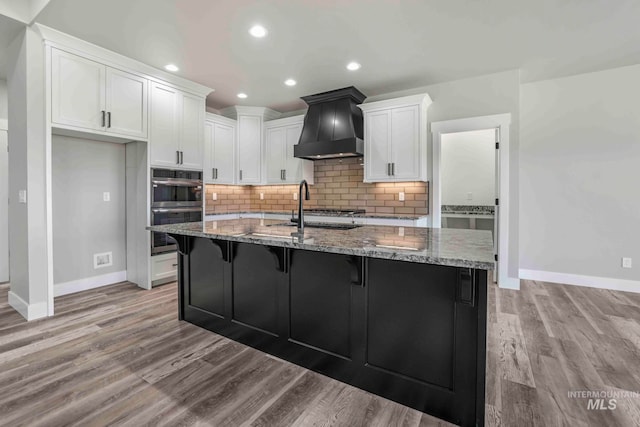 kitchen with a center island with sink, premium range hood, light stone countertops, light hardwood / wood-style flooring, and sink