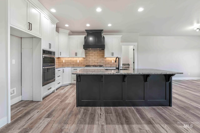 kitchen with appliances with stainless steel finishes, premium range hood, a kitchen island with sink, light stone countertops, and sink